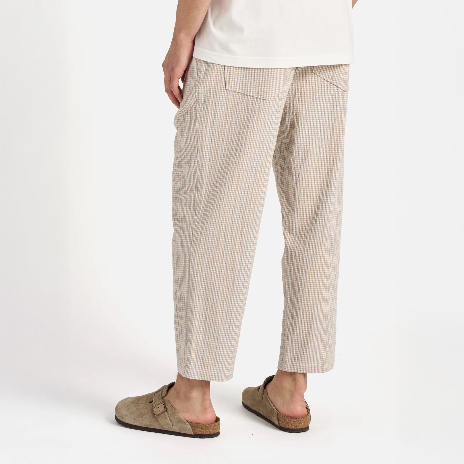A person stands with their back to the camera against a plain white background, wearing Universal Works Ospina Check Cotton High Water Trouser in Ecru/Sand and light brown slip-on sandals.