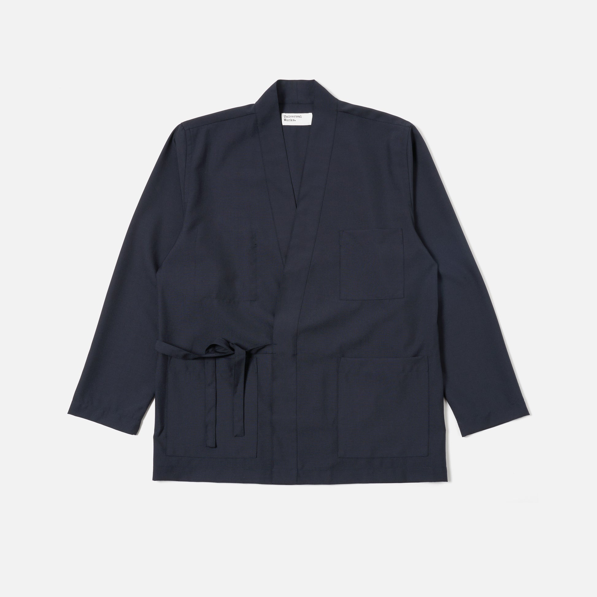 The Universal Works Tropical Suiting Kyoto Work Jacket in navy features long sleeves, a waist tie, and three front pockets, displayed flat on a white background.