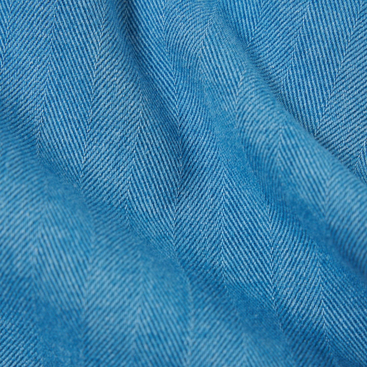 Close-up of the Universal Works Herringbone Recycled Kyoto Work Jacket in Washed Indigo, showing its blue denim fabric with a chevron herringbone pattern and visible diagonal texture.
