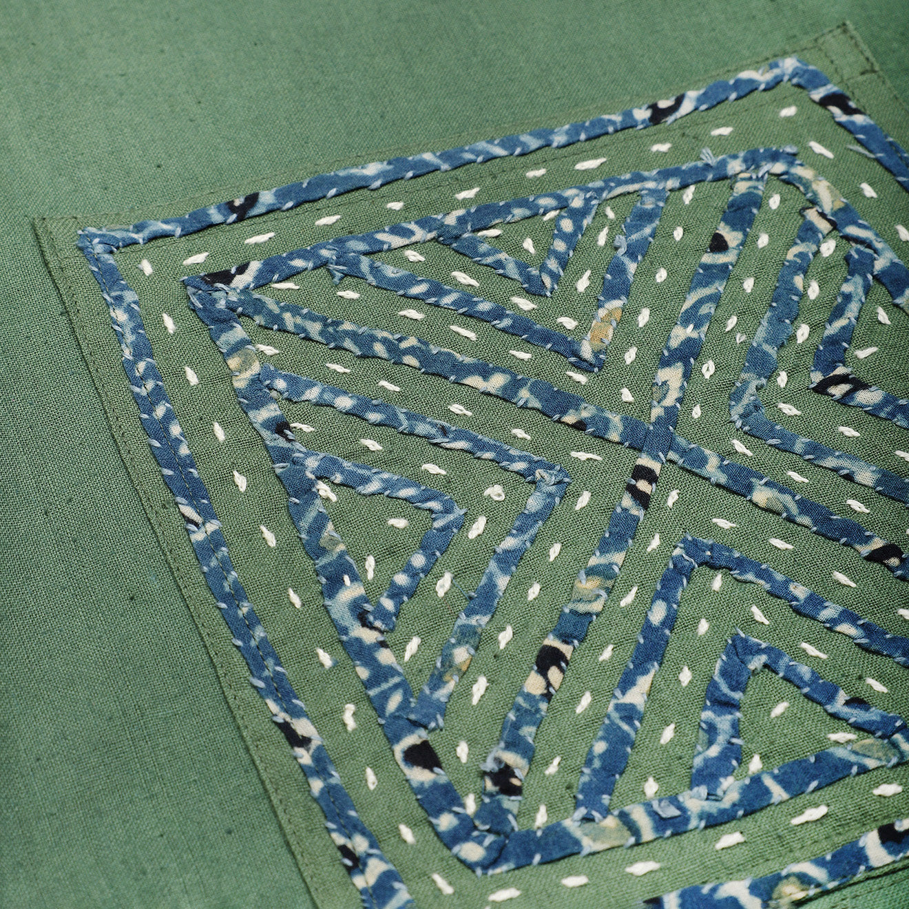 Close-up of Kardo's Bodhi Jacket in Matcha Rabari Applique, featuring green fabric with blue geometric patchwork and white running stitch accents.