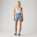 A person with long blonde hair, dressed in a white sleeveless top, Levis 501 Original Short, and blue sneakers stands facing away against a plain light background.