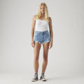 A woman faces forward against a plain light background, wearing a white tank top, Levi’s 501 Original Shorts, crew socks, and black sneakers.