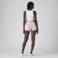A person faces away, wearing a white tank top, Levi’s 501 Original Short in vintage-inspired high-rise light pink denim with frayed edges, and white slip-on shoes against a plain background.