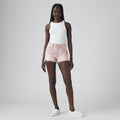A woman stands against a plain background wearing a white sleeveless top, Levis 501 Original Short in vintage-inspired light pink, and white sneakers.