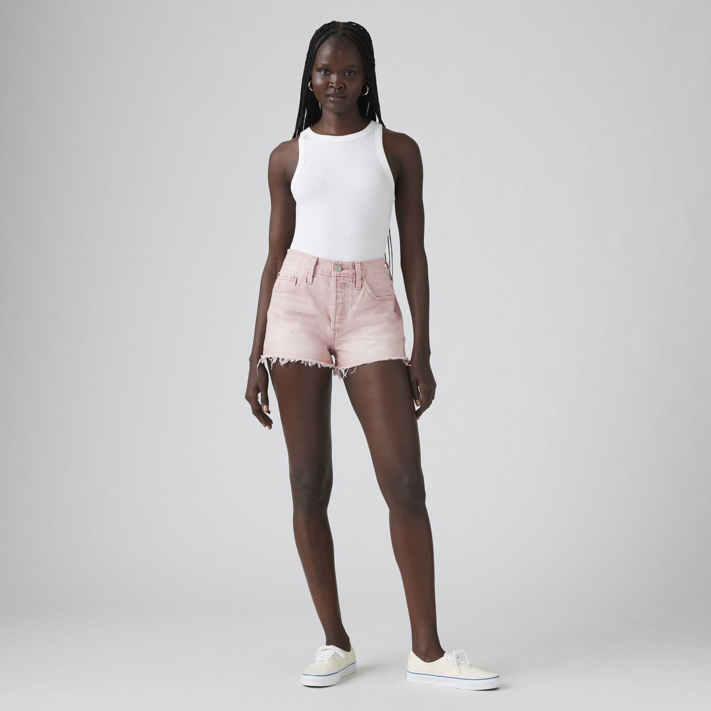 A woman stands against a plain background wearing a white sleeveless top, Levis 501 Original Short in vintage-inspired light pink, and white sneakers.
