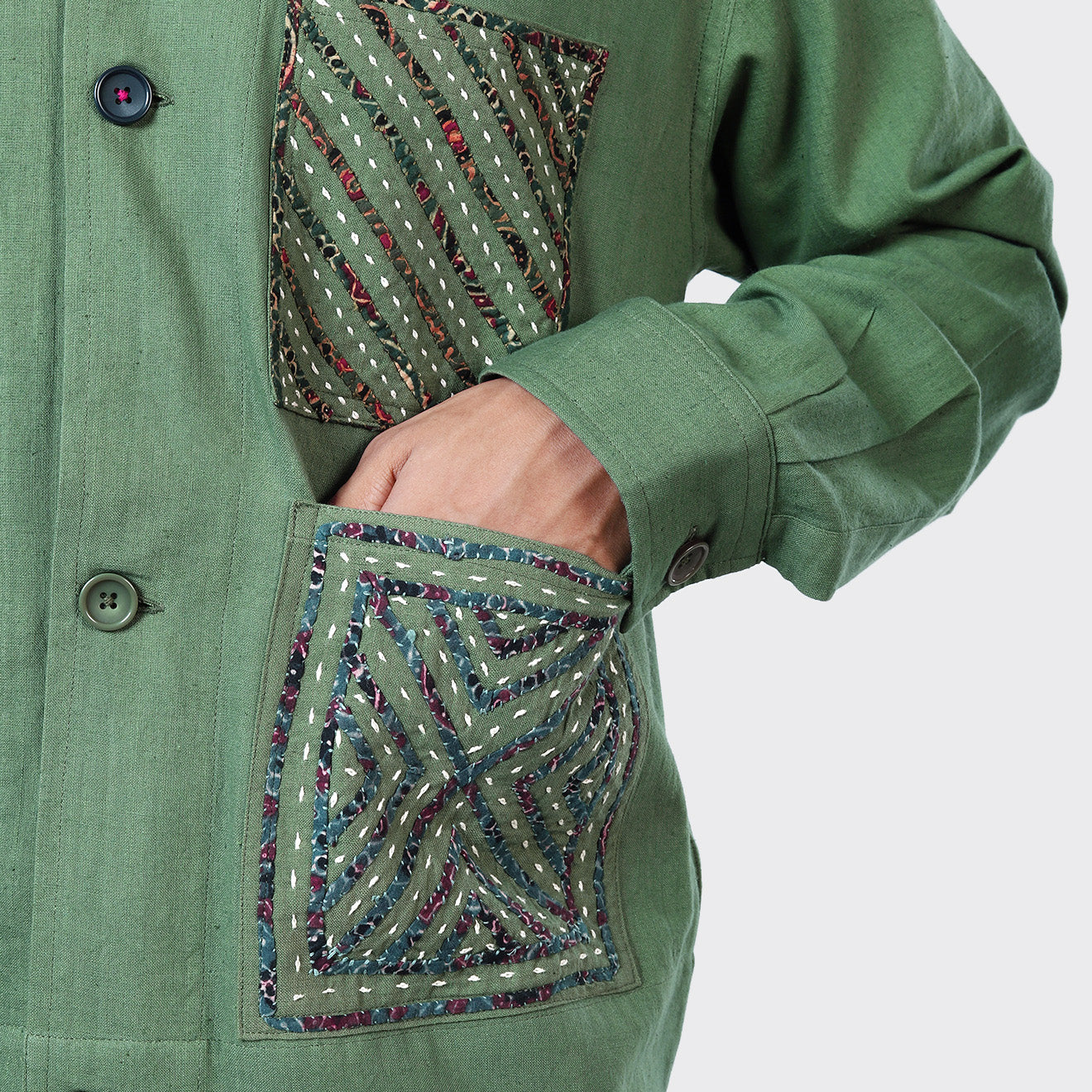A person wearing Kardo’s Bodhi Jacket in Matcha Rabari Applique, featuring patterned embroidery on the chest and hip pockets, places their hand into a lower pocket.