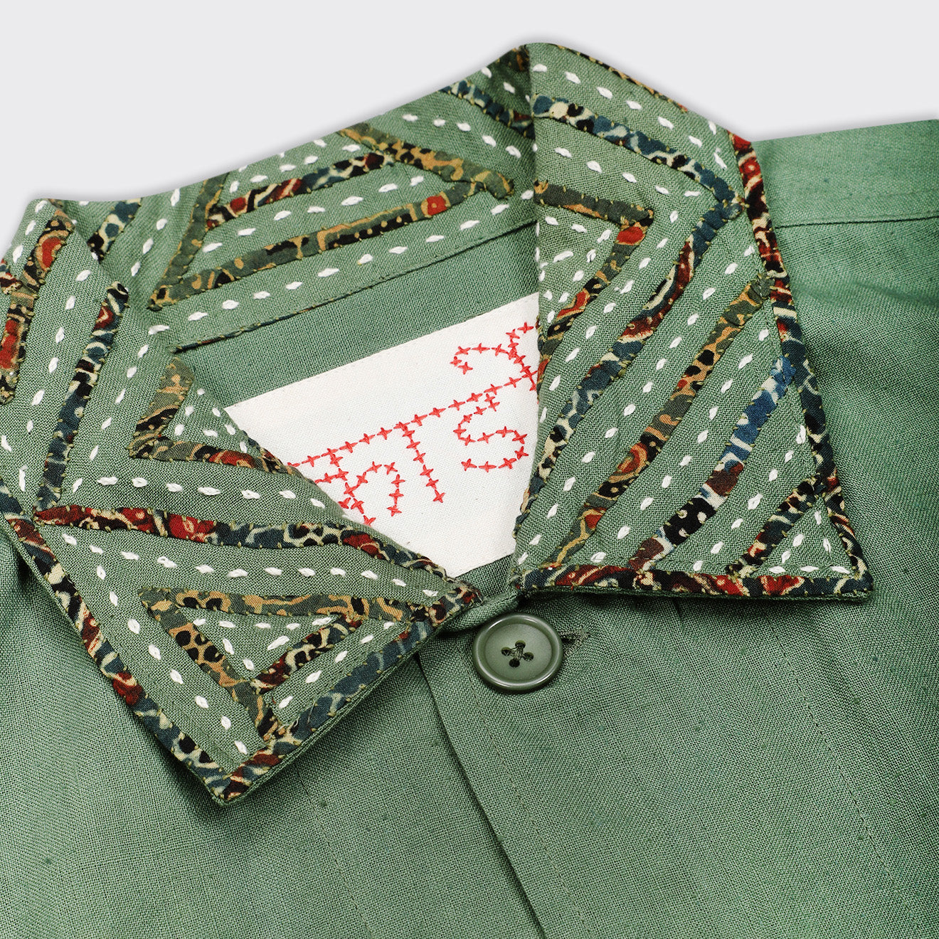A close-up of the Kardo Bodhi Jacket in Matcha Rabari Applique highlights a button, its patterned collar, and a partially visible white label with red stitching.