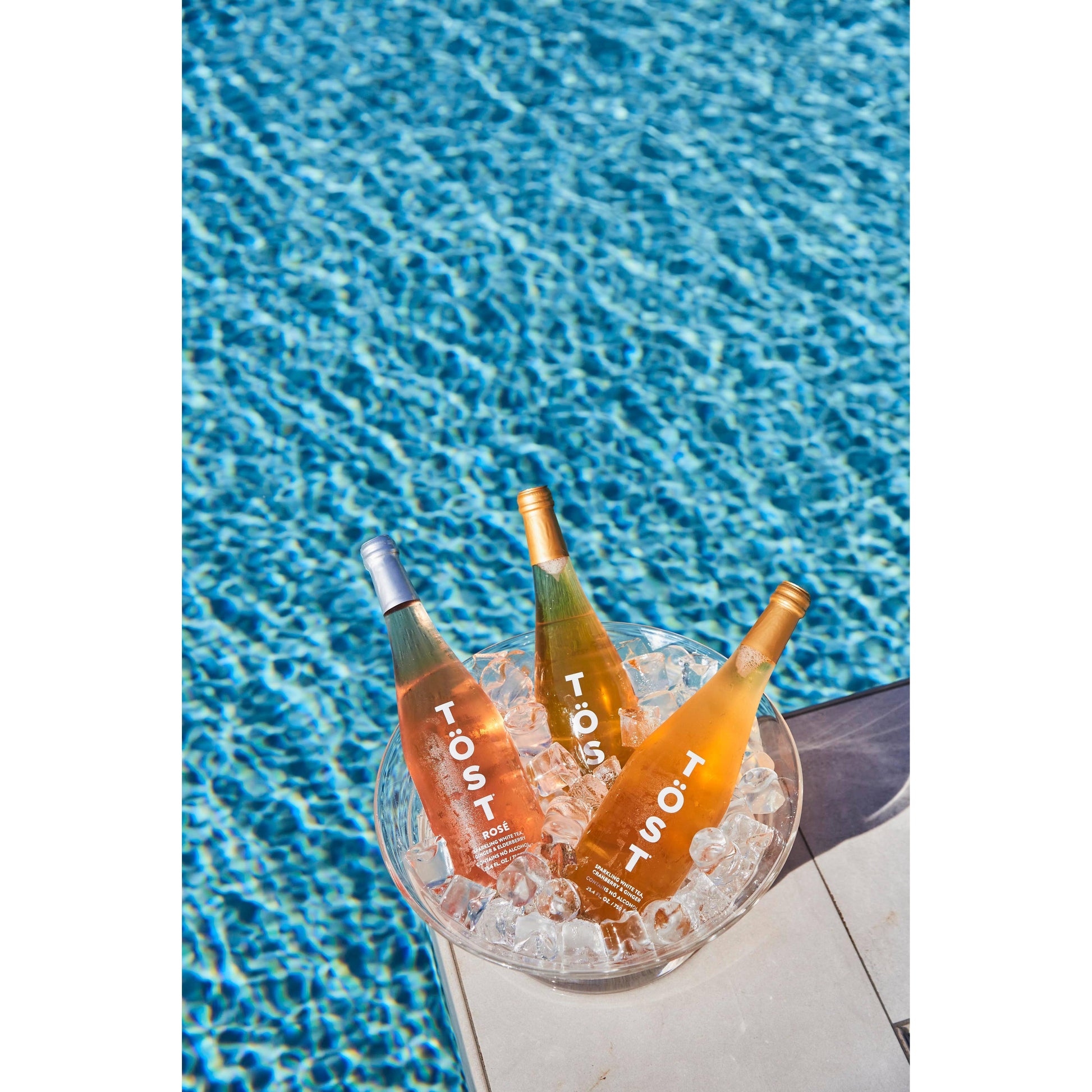 Three bottles of TÖST ROSÉ chilling in a bucket of ice beside a swimming pool with clear blue water.