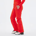 Wearing the Halfdays Alessandra Pant in bright orange with a matching long-sleeve top and white ski boots, a person stands with one hand in their pocket.