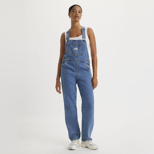 A person stands against a plain background wearing Levis Vintage Overall with adjustable straps, styled with a white tank top and classic white sneakers.