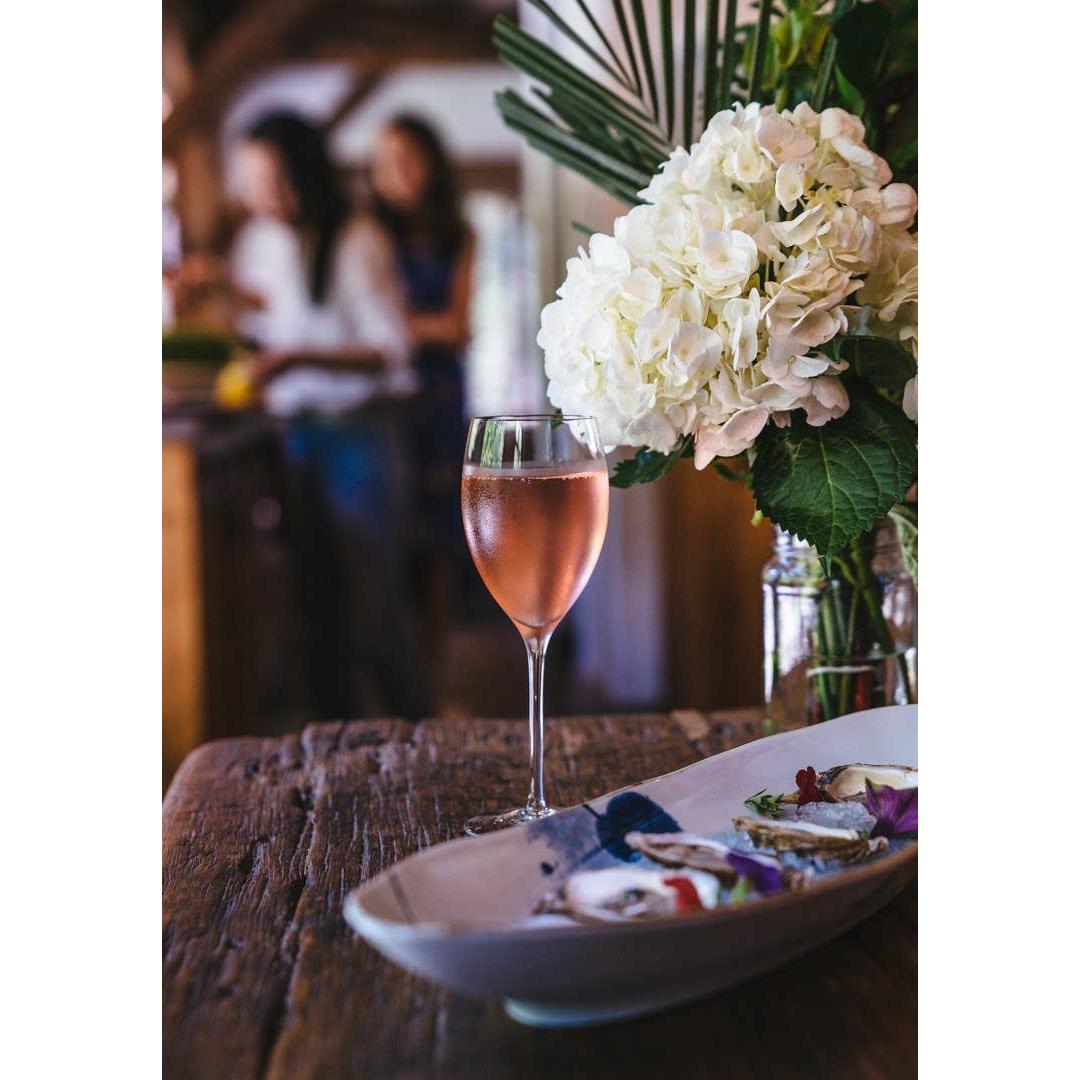 A glass of TÖST ROSÉ by TÖST Beverages next to a plate of food, with a bouquet of hydrangeas and blurry figures in the background.