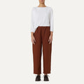 Against a neutral backdrop, a person wears Labo Art's Panta Vela Pant in rust, complete with an elastic waistband and a white long-sleeve top. Dark shoes complete this effortlessly stylish look.