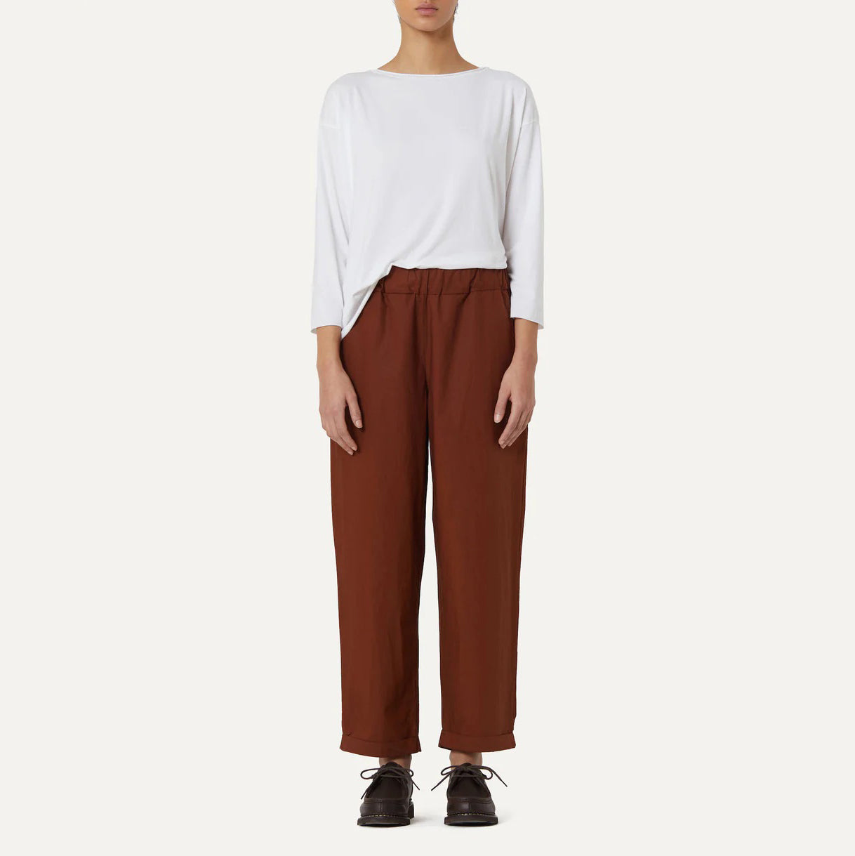 Against a neutral backdrop, a person wears Labo Art's Panta Vela Pant in rust, complete with an elastic waistband and a white long-sleeve top. Dark shoes complete this effortlessly stylish look.