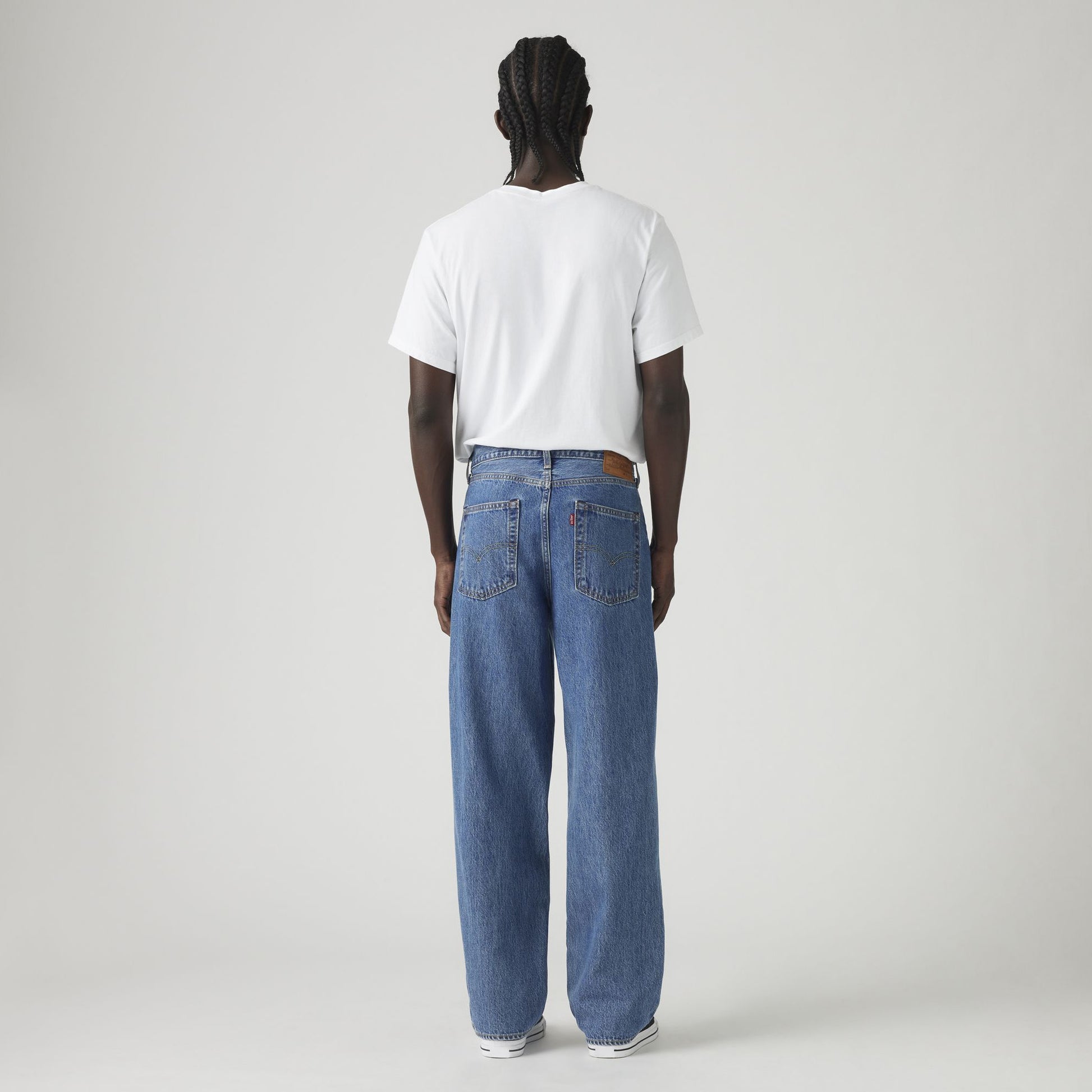 A person stands facing away, wearing a white T-shirt, Levis 578 Baggy loose blue jeans with a baggy silhouette, and sneakers, against a plain light background.