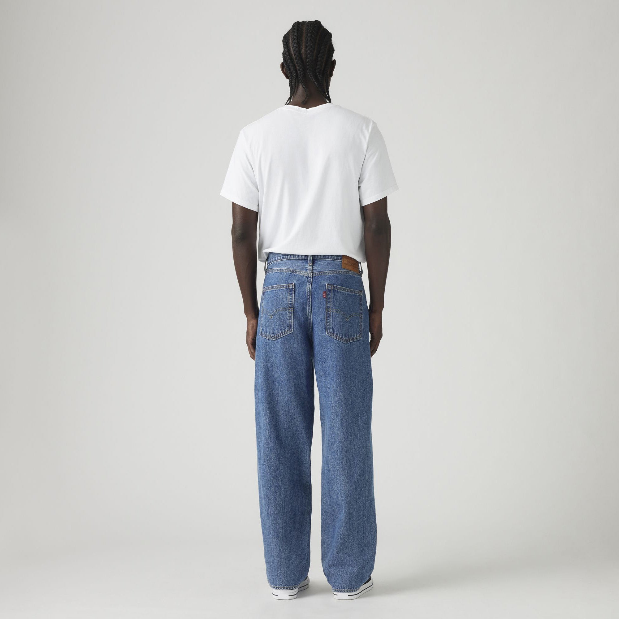 A person stands facing away, wearing a white T-shirt, Levis 578 Baggy loose blue jeans with a baggy silhouette, and sneakers, against a plain light background.