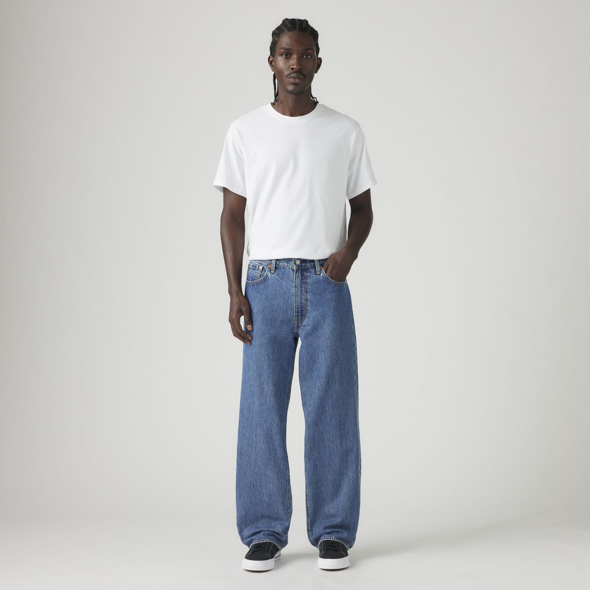 A man faces forward against a plain light background, wearing a white t-shirt, black sneakers, and Levis 578 Baggy blue jeans in a loose silhouette.