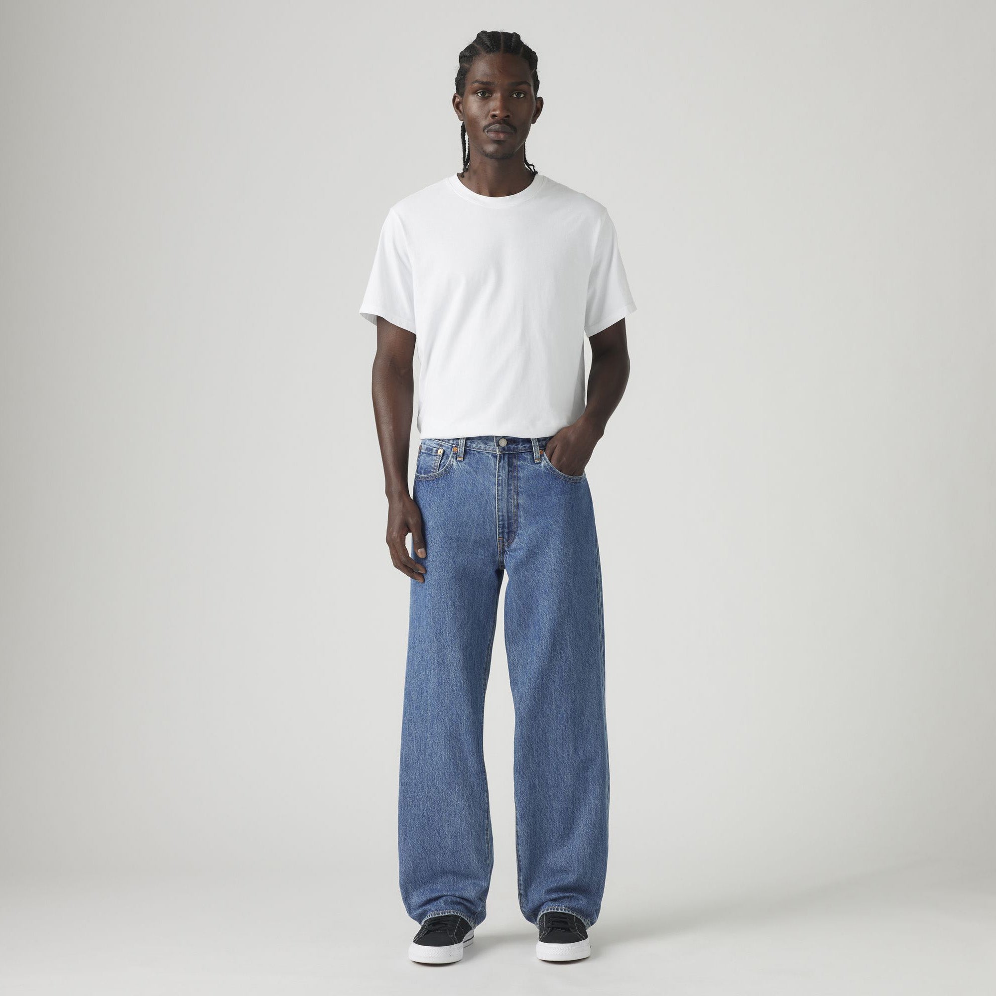A man faces forward against a plain light background, wearing a white t-shirt, black sneakers, and Levis 578 Baggy blue jeans in a loose silhouette.