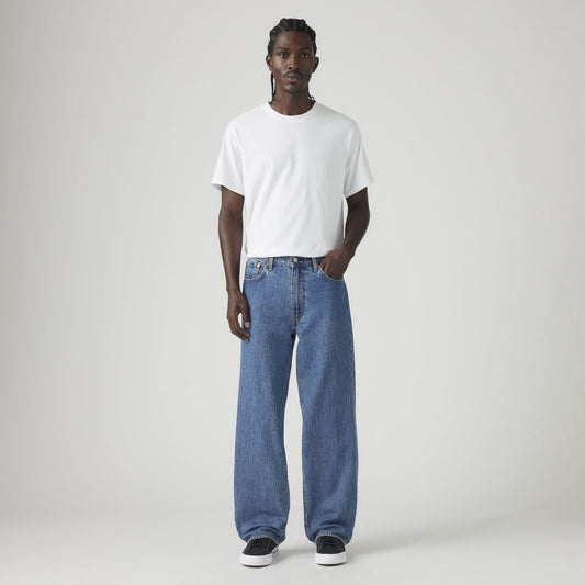 A man faces forward against a plain light background, wearing a white t-shirt, black sneakers, and Levis 578 Baggy blue jeans in a loose silhouette.