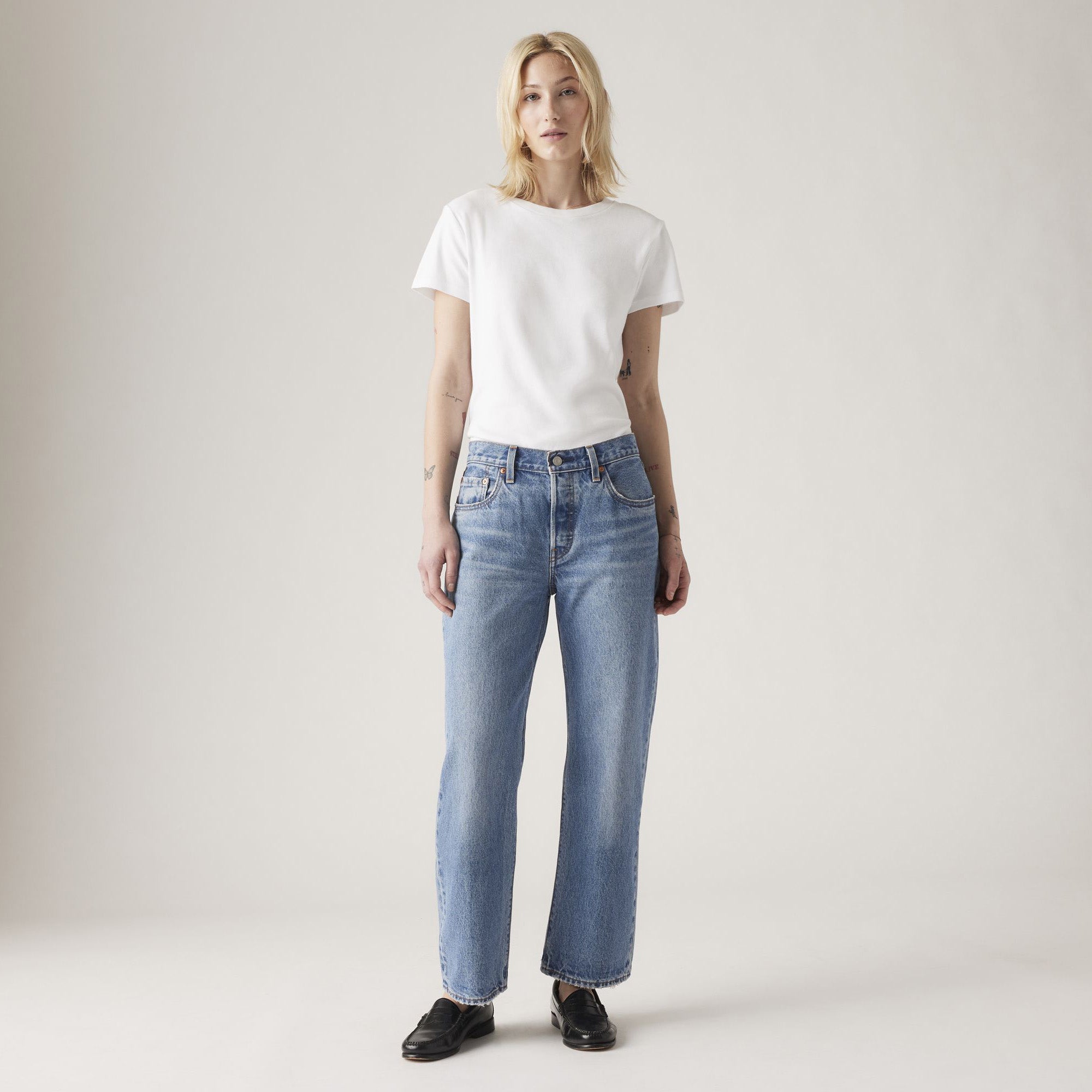 A person stands against a plain background wearing a white t-shirt, black shoes, and Levis 501 90s Ankle jeans in the Sweetest Taboo light blue wash.