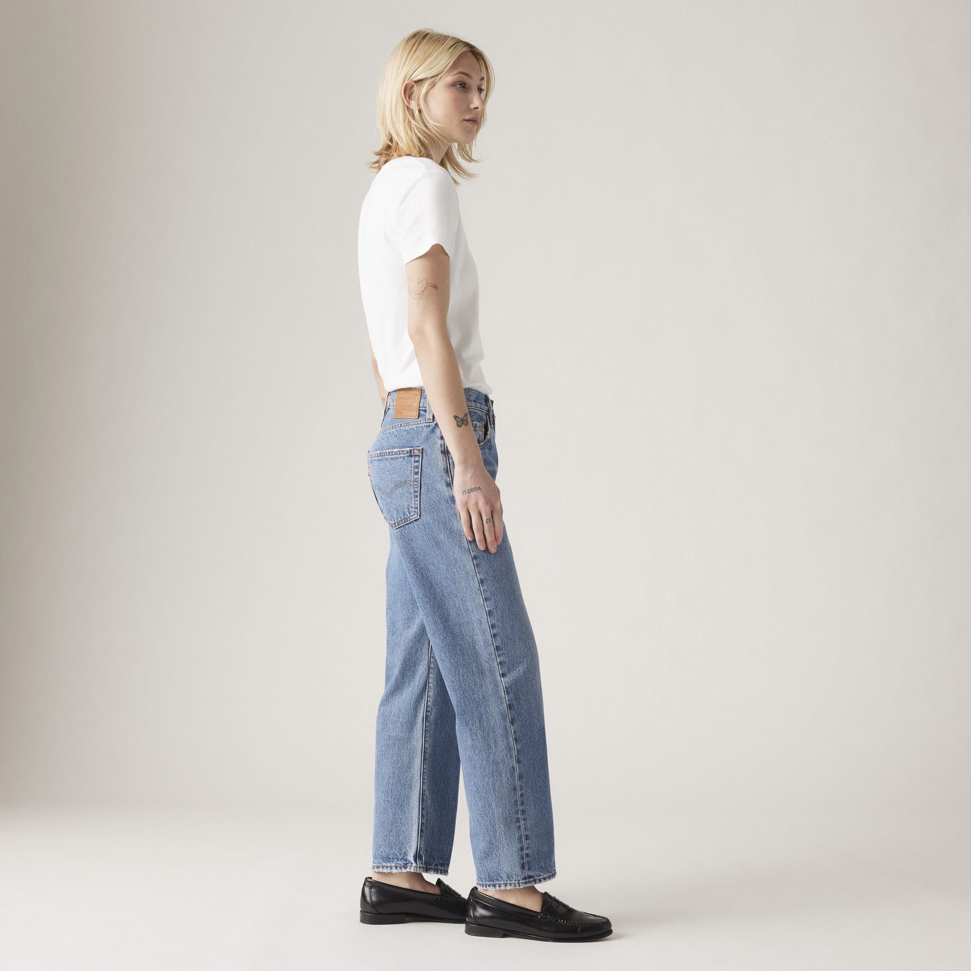 A person stands sideways in a white t-shirt, Levis 501 90s Ankle jeans in Sweetest Taboo blue wash, and black loafers against a plain light background.