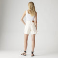 A person with long blonde hair, wearing a white tank top and Levis High Baggy Short in cream, paired with black sandals, stands facing away in a minimalist studio.