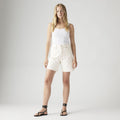 A woman stands against a plain background wearing a white tank top, Levis High Baggy Short, and black strappy sandals.