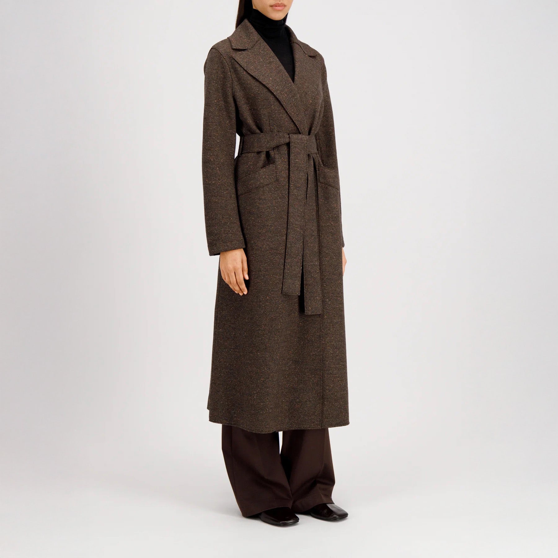 A woman wears the Harris Wharf W Long Maxi Coat Donegal—a long, brown belted coat—over dark trousers and black shoes, standing against a plain light background.