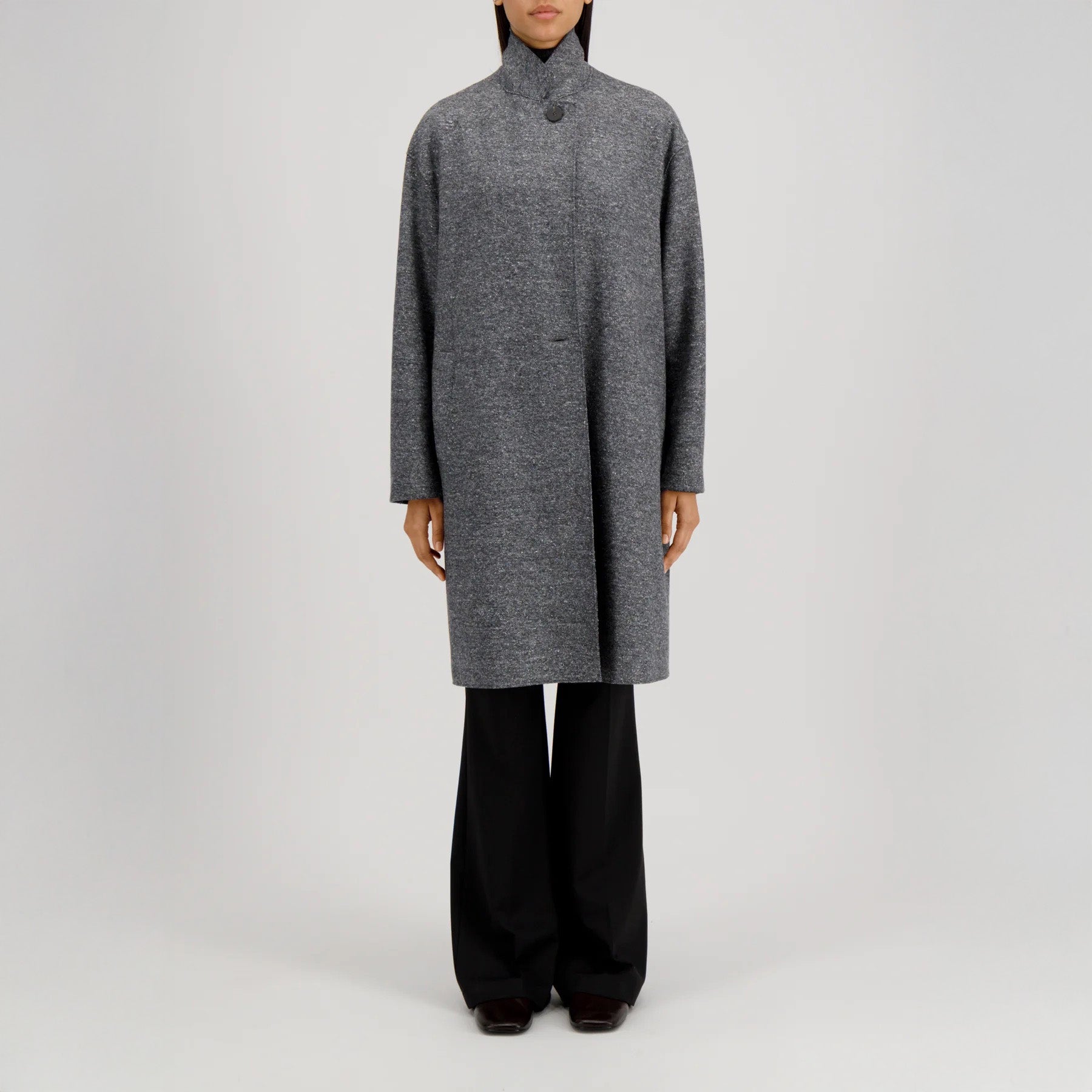 A person stands against a plain background wearing the Harris Wharf W Oversized Coat Donegal in grey, styled over black pants and dark shoes.