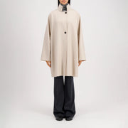 A model wears the Harris Wharf W Raglan Cape in pressed wool, beige and knee-length with buttons, over dark wide-leg pants and black shoes, standing against a plain white background.