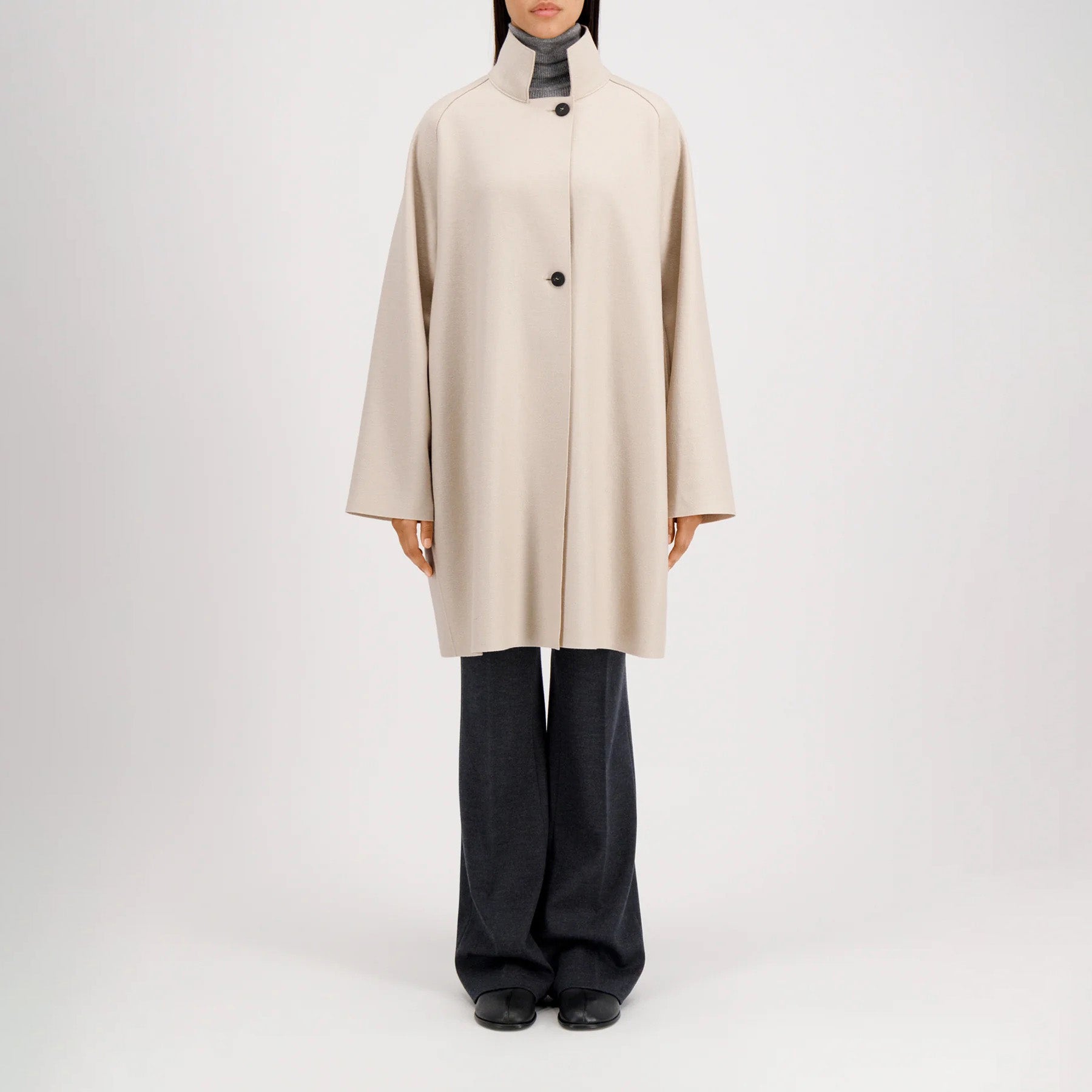 A model wears the Harris Wharf W Raglan Cape in pressed wool, beige and knee-length with buttons, over dark wide-leg pants and black shoes, standing against a plain white background.