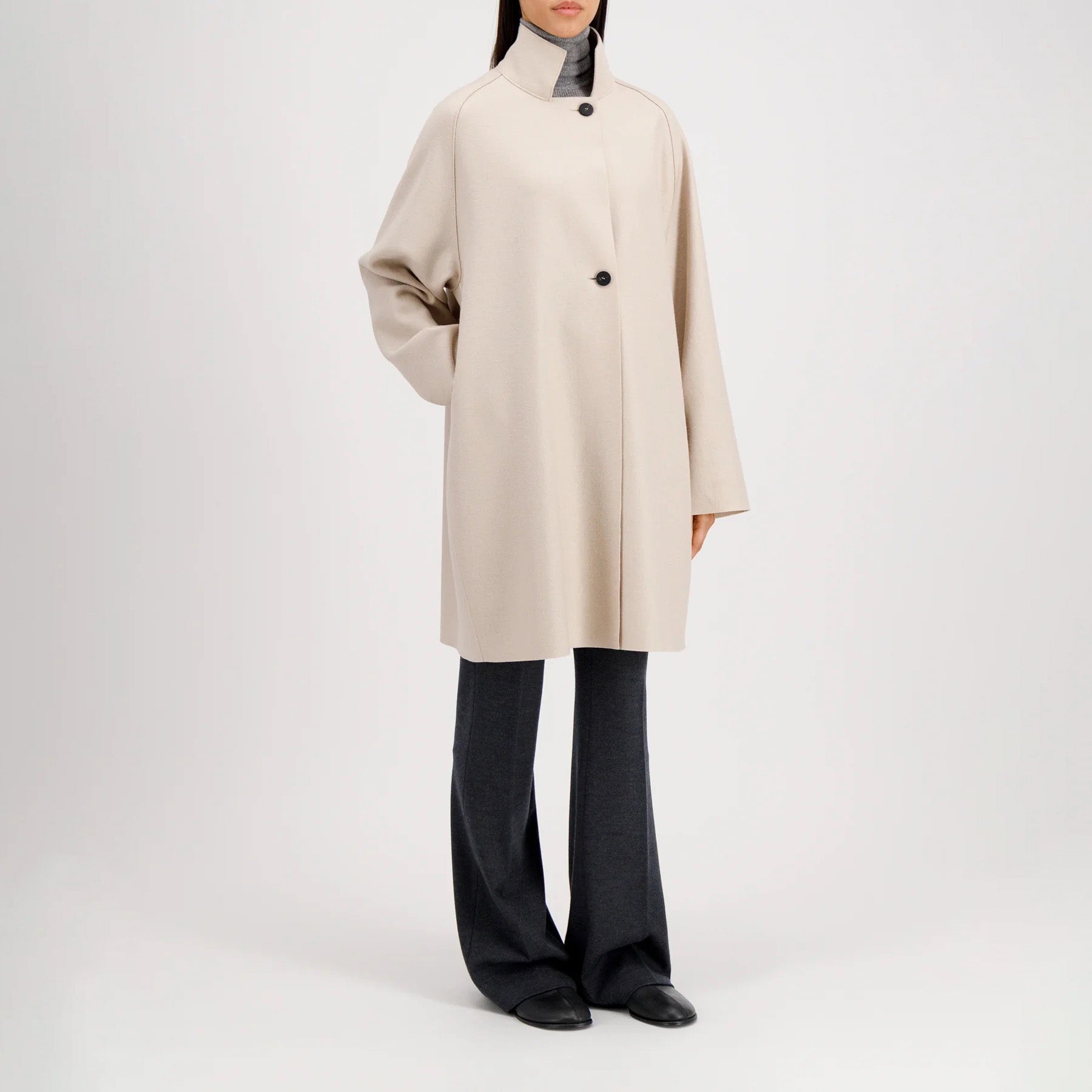 A person stands against a plain white background wearing the Harris Wharf W Raglan Cape in light beige pressed wool, featuring two buttons, paired with dark gray pants and black shoes. This elegant, knee-length cape is made in Italy for timeless style.