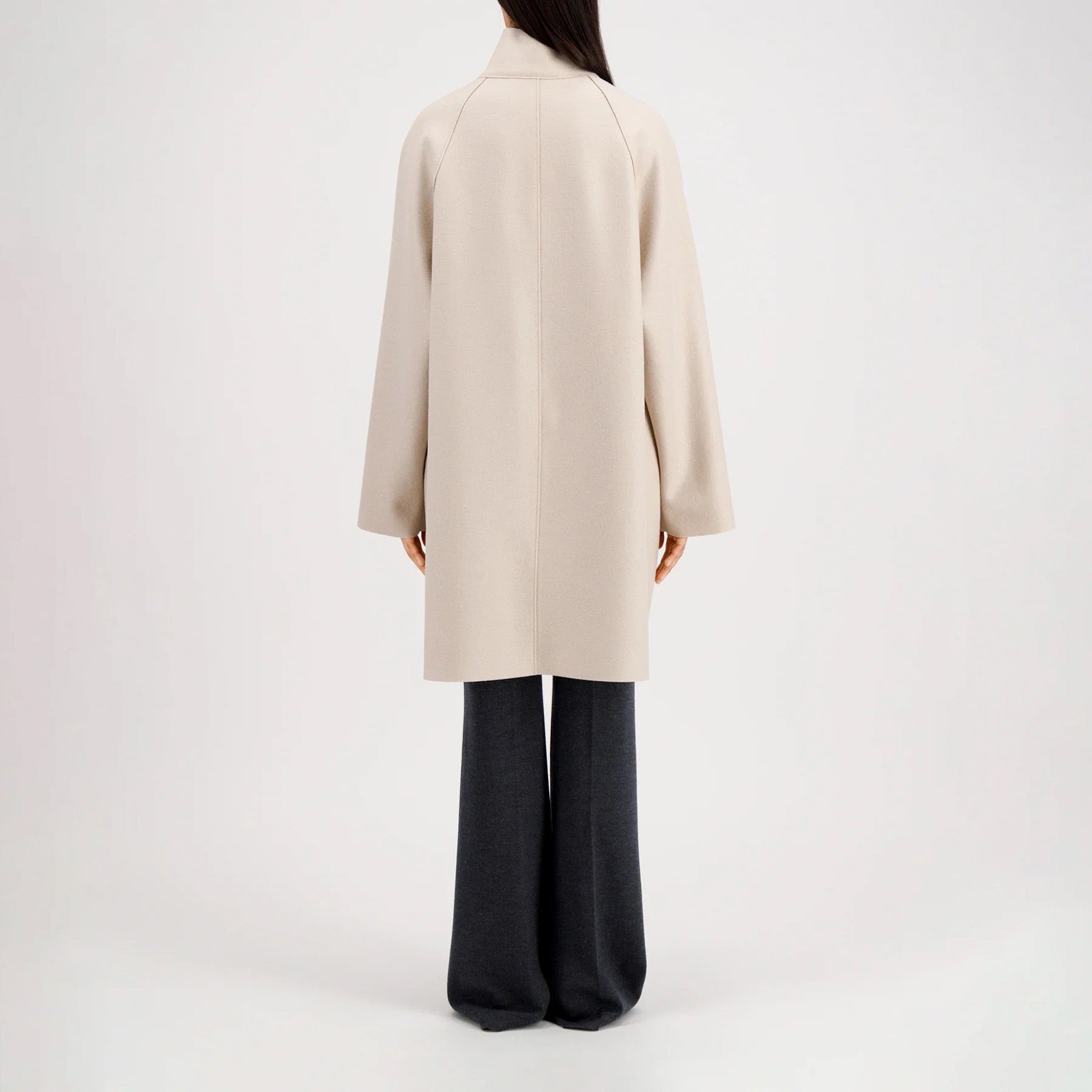 A person stands facing away, wearing the Harris Wharf W Raglan Cape in pressed wool with dark wide-leg pants against a plain light background.