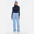 A person is wearing a black and blue long-sleeve top, light blue Halfdays Alessandra Pant with a ski fit, a core classic blue beanie, and white shoes while standing against a plain white background.