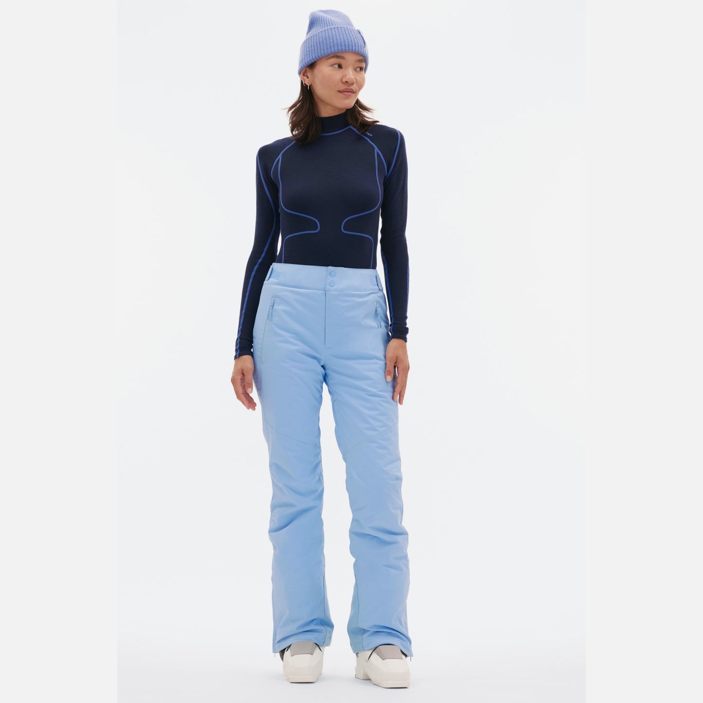 A person is wearing a black and blue long-sleeve top, light blue Halfdays Alessandra Pant with a ski fit, a core classic blue beanie, and white shoes while standing against a plain white background.