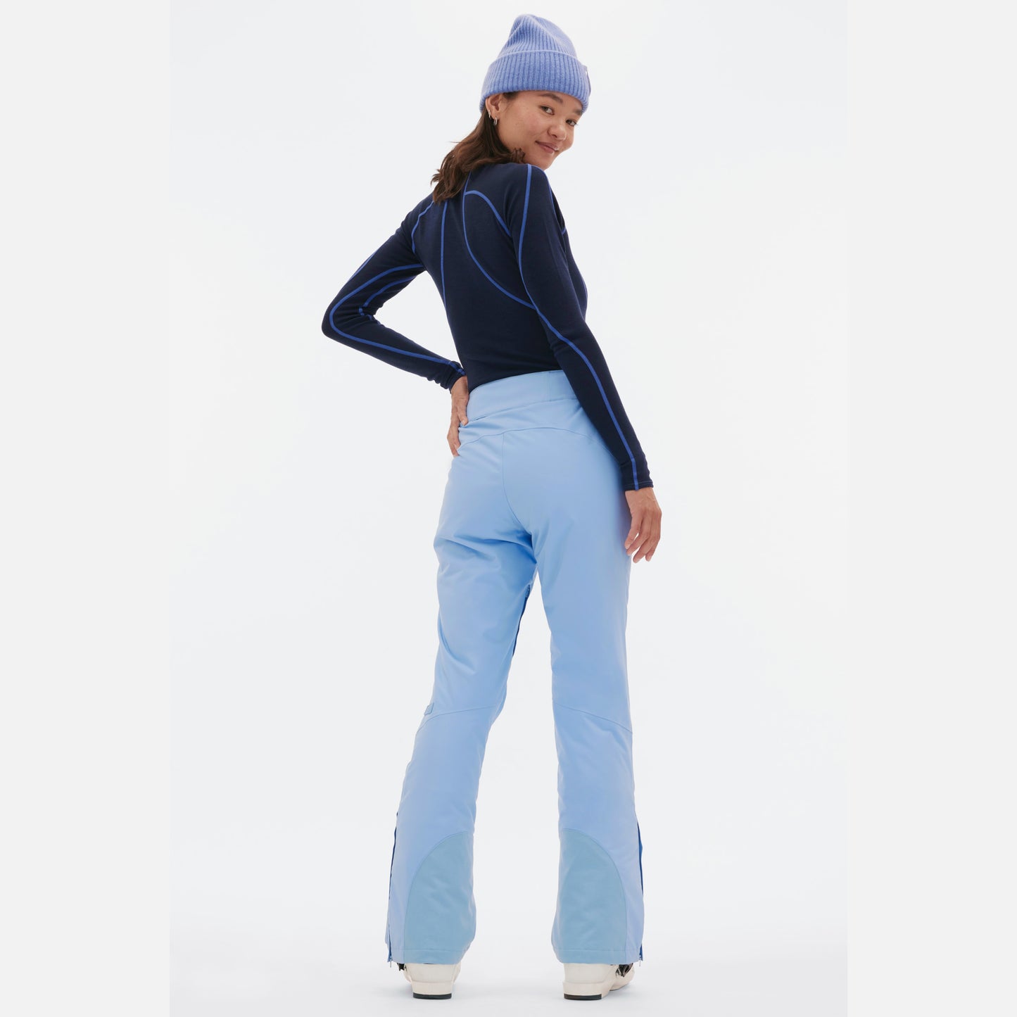 A person wearing a dark blue long-sleeve top, the light blue Halfdays Alessandra Pant, and a matching knit beanie, stands smiling while looking back.