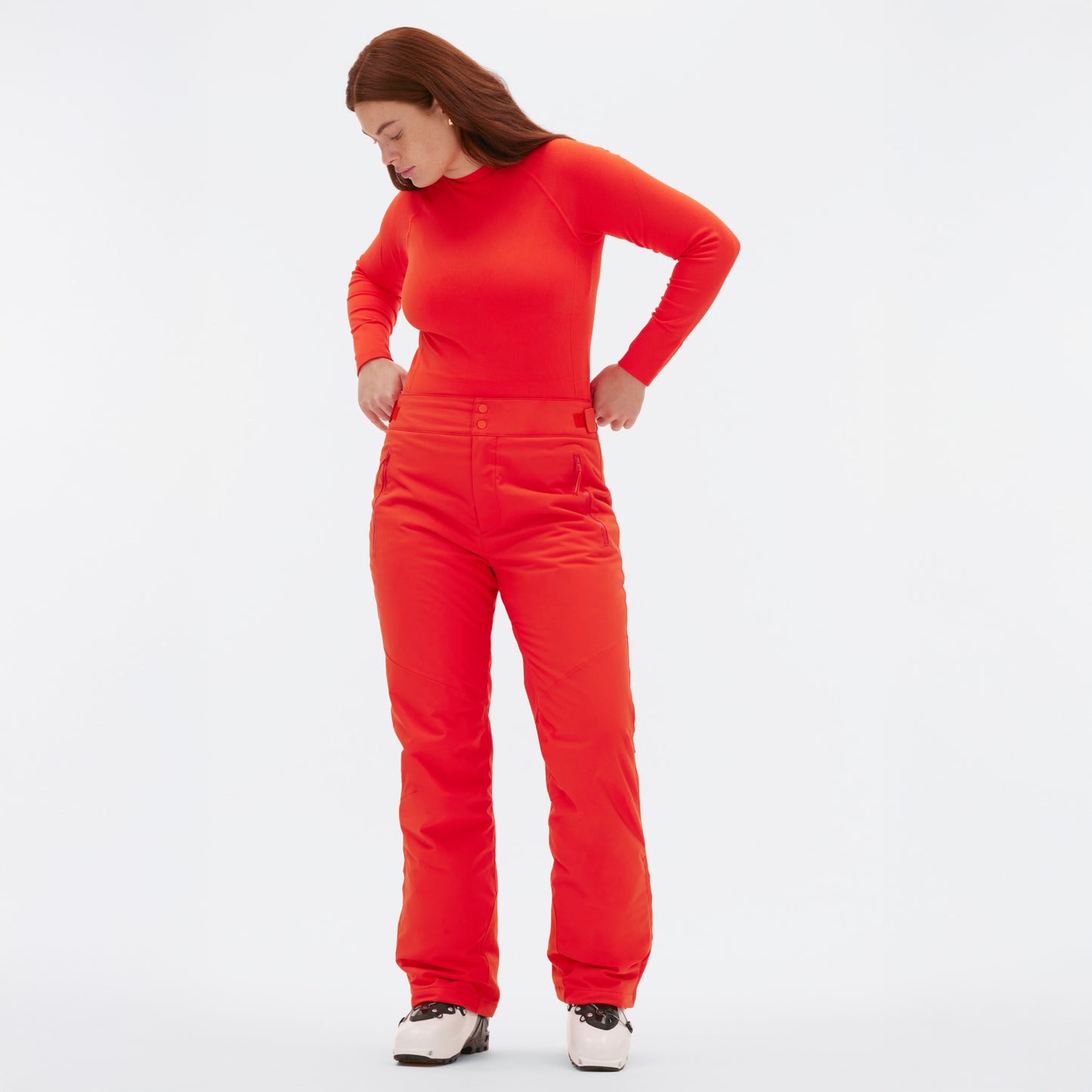 A person faces slightly downward, wearing the Halfdays Alessandra Pant with a matching red long-sleeve top and white ski boots against a plain white background.