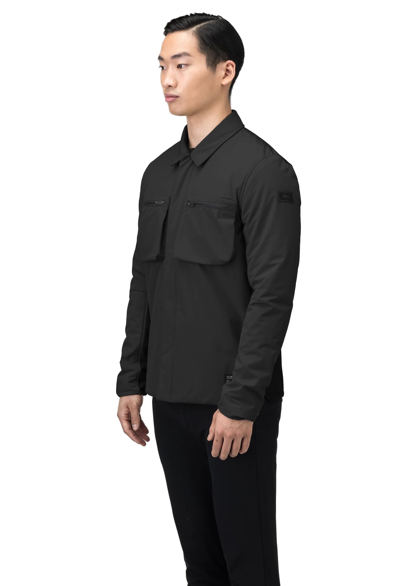 A person wearing the Ander Men's Mid Layer Shirt by Nobis, a black button-up featuring chest pockets with PrimaLoft Gold Insulation, stands against a white background.