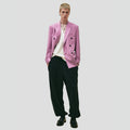 Dressed in Seven Gauge's Double Breast Blazer in a light pink hue over a white shirt, paired with black wide-leg pants and black shoes, the individual exudes a modern yet classic vibe against a plain background.