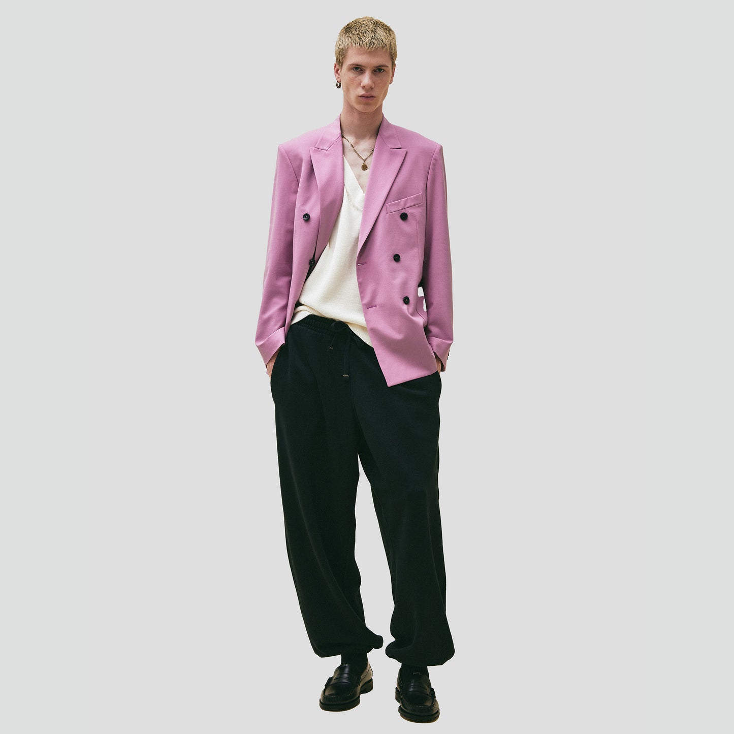 Dressed in Seven Gauge's Double Breast Blazer in a light pink hue over a white shirt, paired with black wide-leg pants and black shoes, the individual exudes a modern yet classic vibe against a plain background.