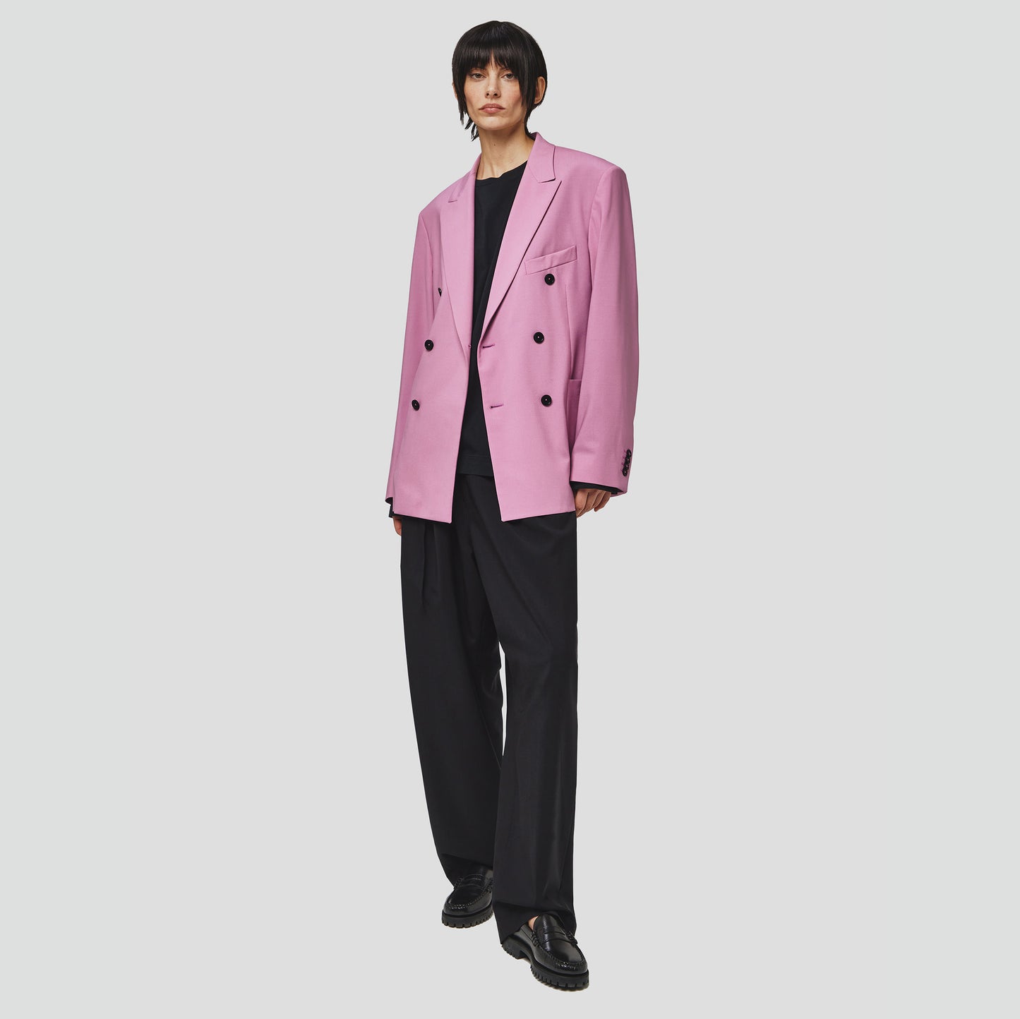 A person stands confidently in a tropical wool double breast blazer by Seven Gauge, paired with a black shirt and black wide-leg trousers against a plain backdrop.