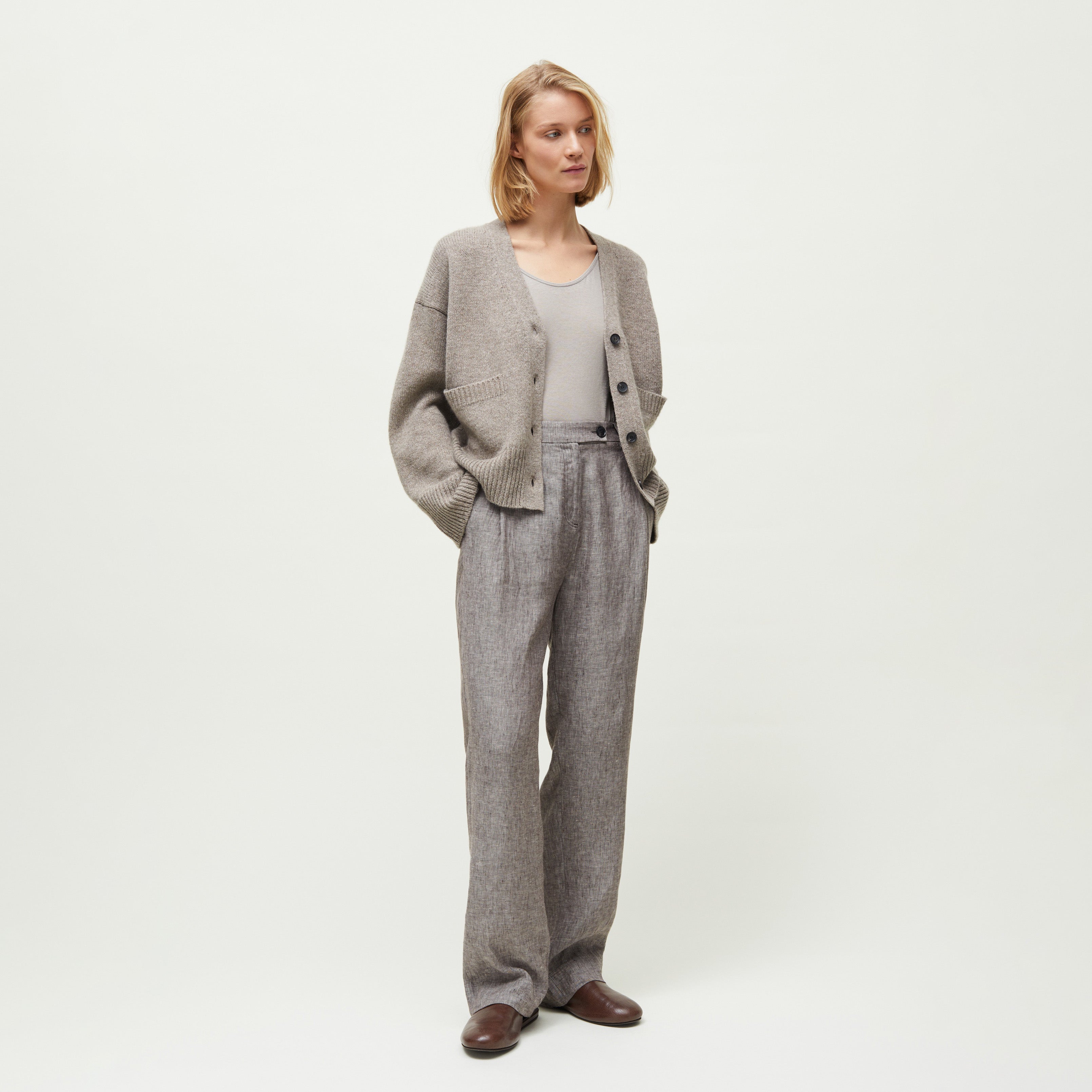 A woman stands against a plain background wearing a grey cardigan, light grey top, Aiayu's Balder Linen Pant inspired by menswear styles, and brown shoes, with her hands in her pockets.