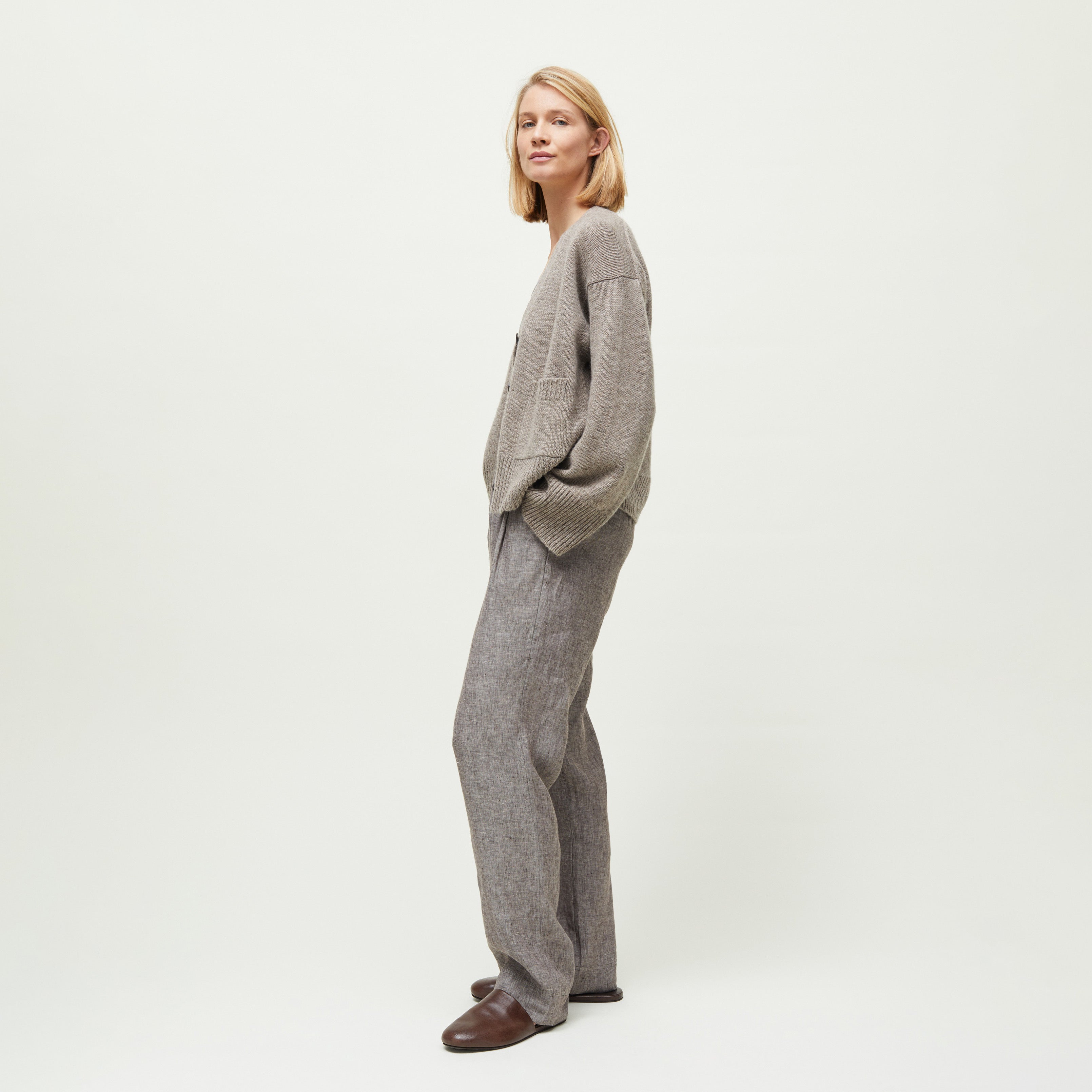 A woman stands sideways against a plain white background, wearing a loose beige sweater, brown shoes, and the Aiayu Balder Linen Pant: wide-leg gray suit trousers inspired by menswear styles.