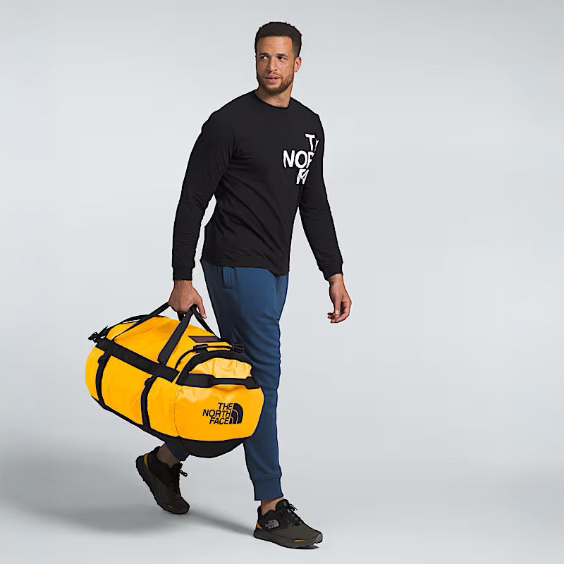 A man in a black long-sleeve shirt and blue pants carries The North Face Base Camp Duffel L in Summit Gold, renowned for its rugged durability, against a plain light background.