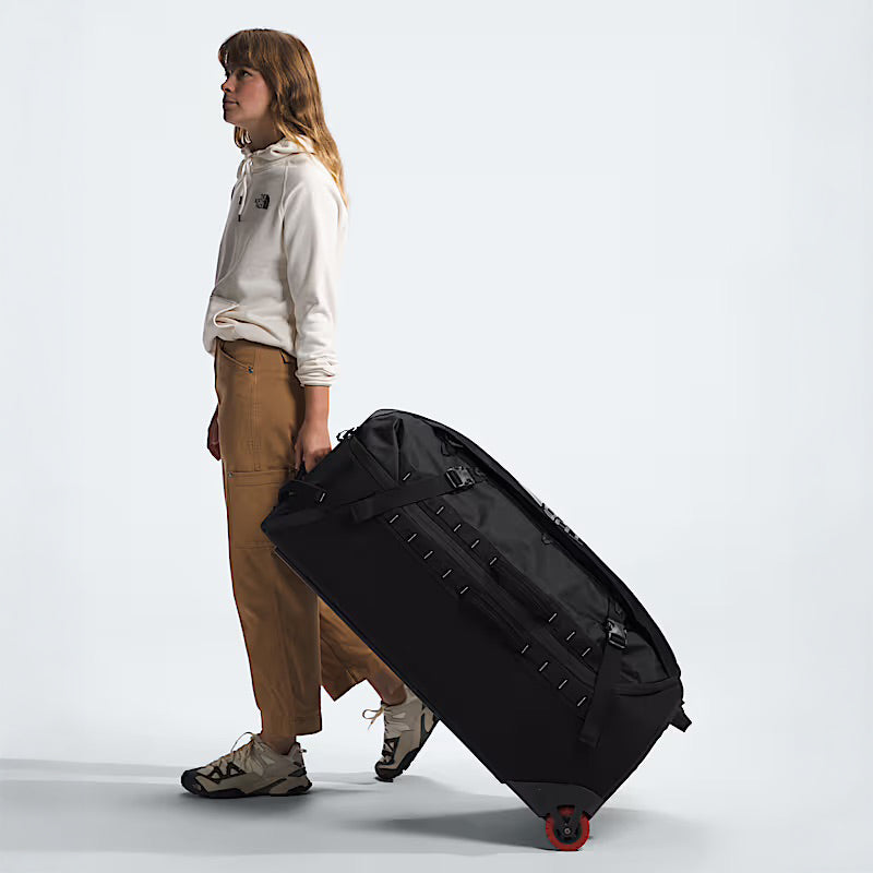 A person in a light hoodie, tan pants, and sneakers pulls The North Face Base Camp Rolling Thunder 36 Luggage—made with recycled fabric and a hard-shell chassis—against a plain light background.
