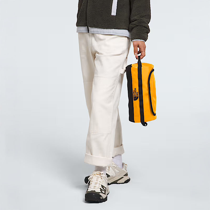 A person wearing off-white pants, white sneakers, and a dark jacket holds The North Face Base Camp Travel Canister in Summit Gold against a plain light gray background.