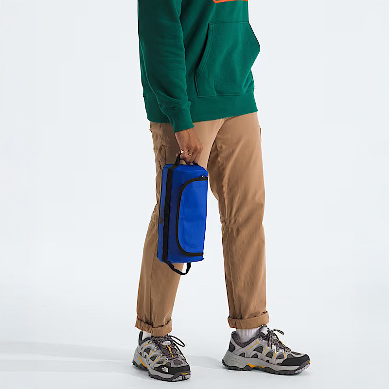 A person wearing a green hoodie, tan pants, and sneakers holds The North Face Base Camp Travel Canister L in Blue/TNF Black—made with durable water-repellent recycled TPE laminate—by its handle against a plain background.