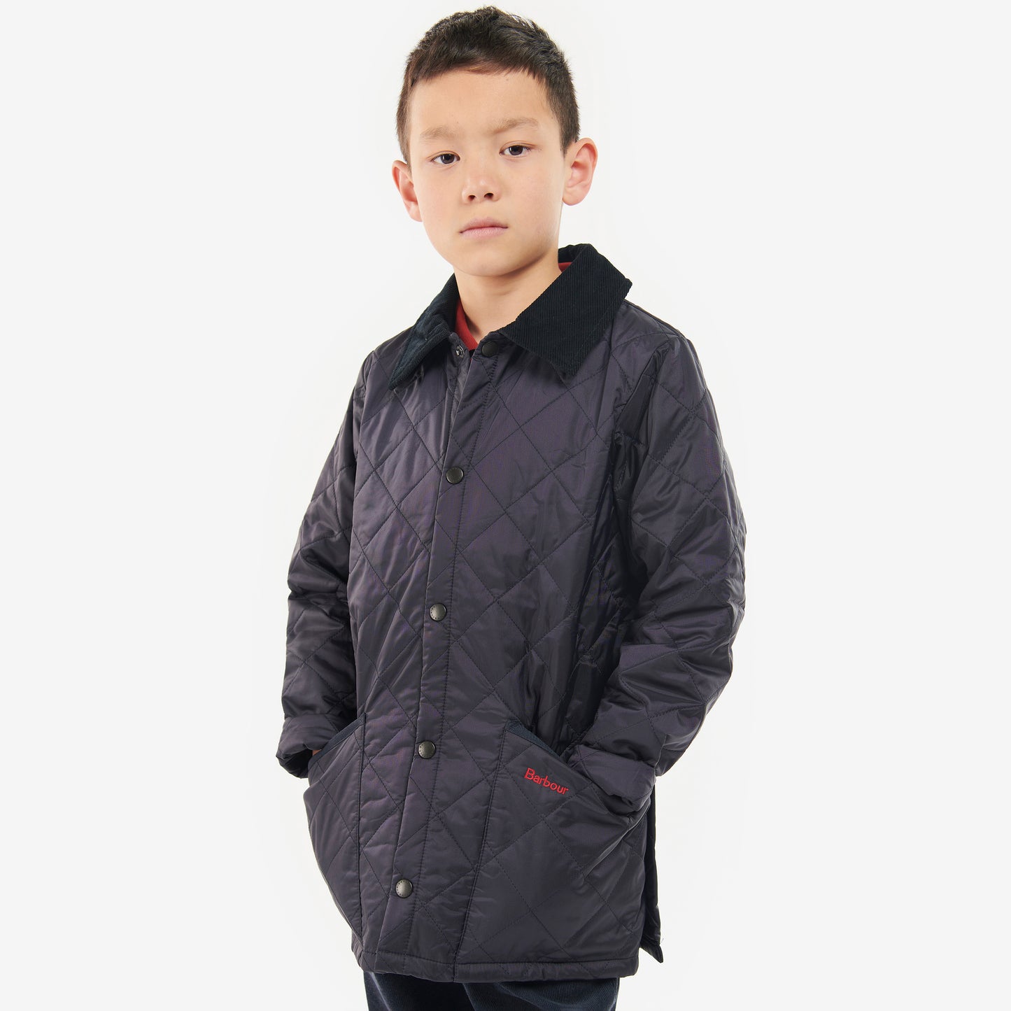 A young boy stands against a white background, hands in the pockets of his Barbour Boys Liddesdale Quilted Jacket. The quilted design and needlecord collar give it charm, and it's perfectly tailored for kids.