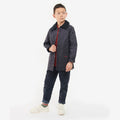 A young boy in a Barbour Boys Liddesdale Quilted Jacket with needlecord collar wears a red shirt, dark pants, and white sneakers. He looks away from the camera.