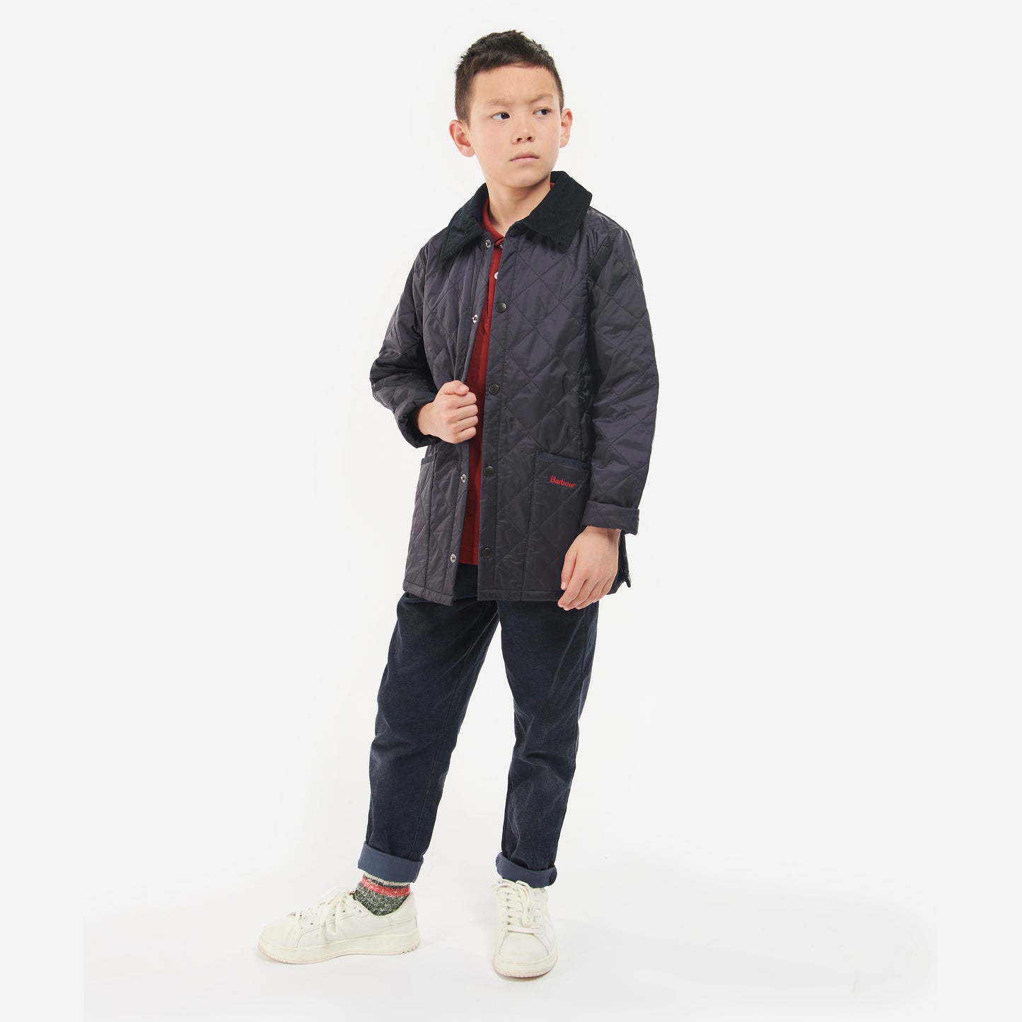 A young boy in a Barbour Boys Liddesdale Quilted Jacket with needlecord collar wears a red shirt, dark pants, and white sneakers. He looks away from the camera.