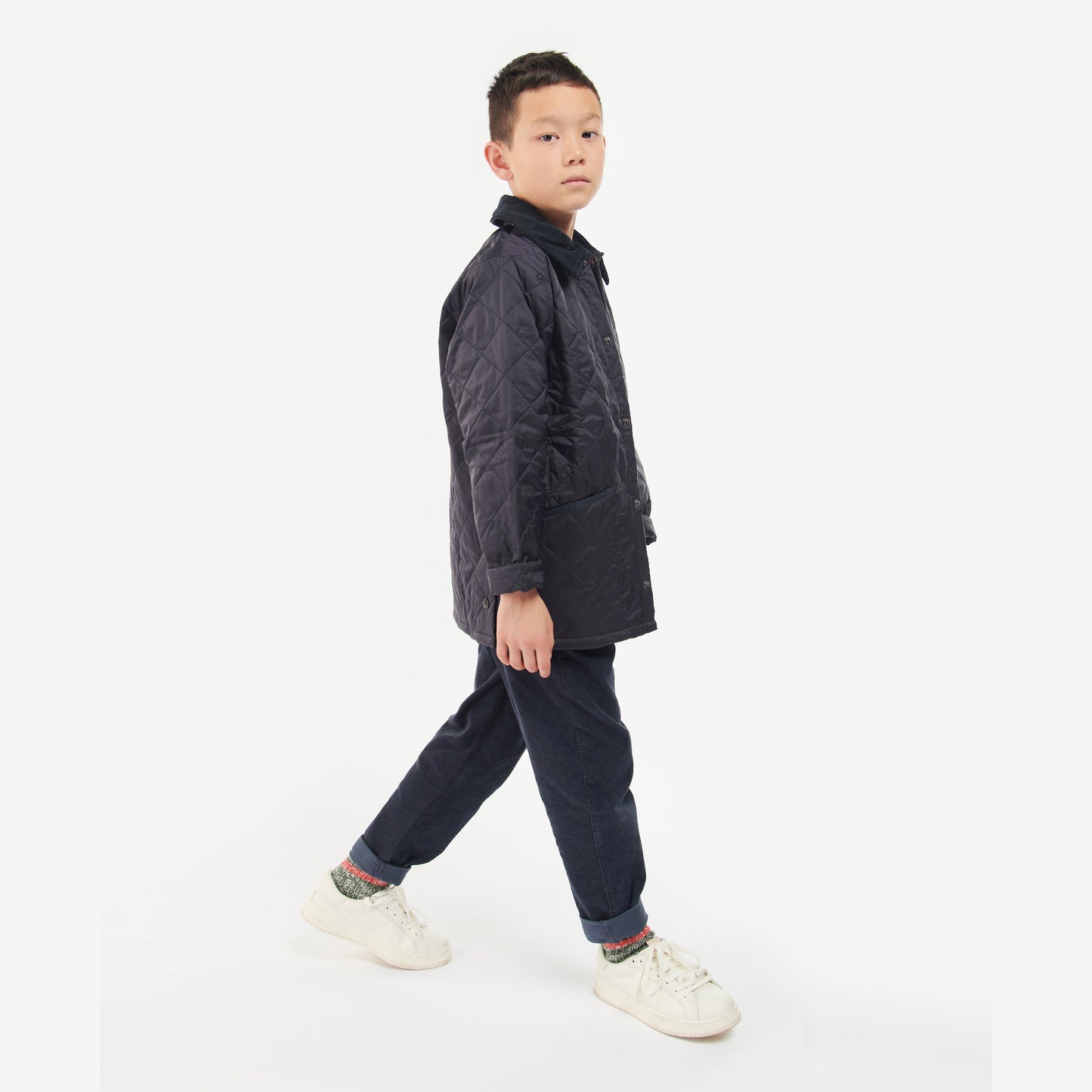 A young boy in a Barbour Boys Liddesdale Quilted Jacket with a needlecord collar, dark pants, and white sneakers stands on a light background, looking forward with one leg slightly raised.